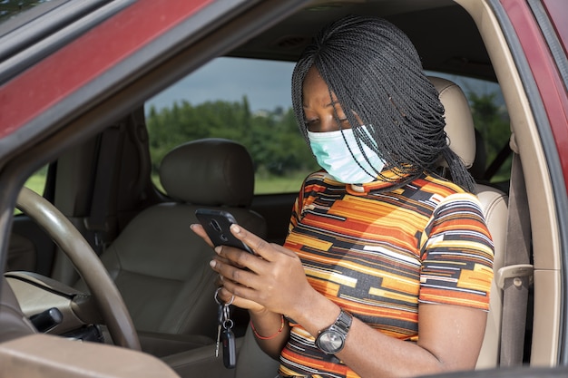 Black woman using her phone while sitting in a car, wearing a face mask - the new normal concept