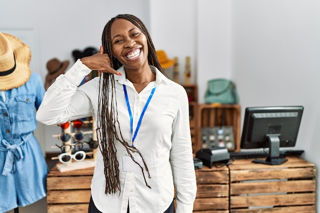 Black woman with braids working as manager at retail boutique smiling doing phone gesture with hand and fingers like talking on the telephone. communicating concepts.