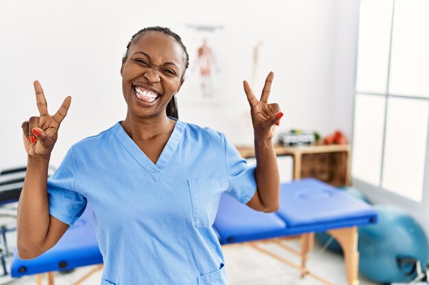 통증 회복 클리닉에서 일하는 흑인 여성은 승리의 표시를 하는 두 손의 손가락을 내보이며 혀를 내밀고 웃고 있습니다. 두 번째.