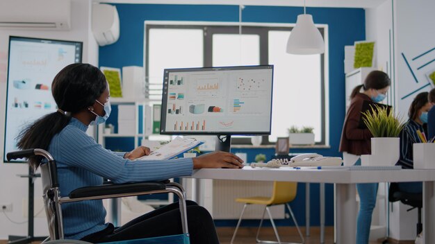 Black woman with disabilities and protection face masks working on computer in new normal workplace during pandemic. Multiethnic team in business financial office checking reports, analysing datas