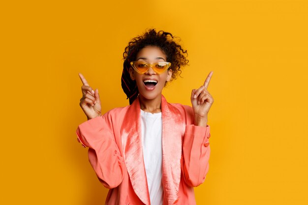Black woman with funny face pointing up. Wearing casual pink jacket. Yellow background .