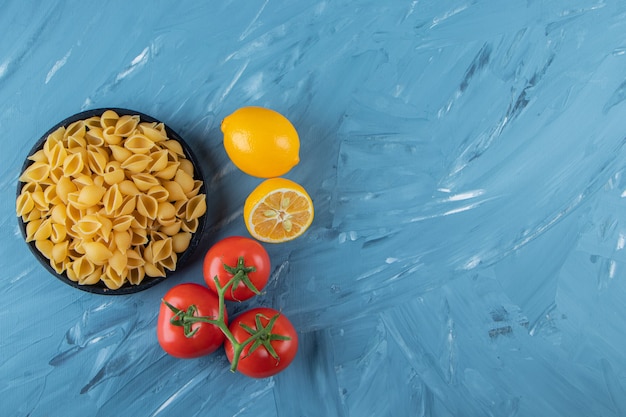 A black wooden plate of raw pasta with lemon and fresh red tomatoes. 