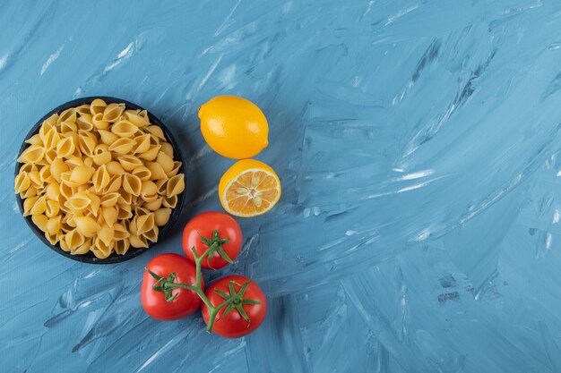 Черная деревянная тарелка сырых макарон с лимоном и свежими красными помидорами.