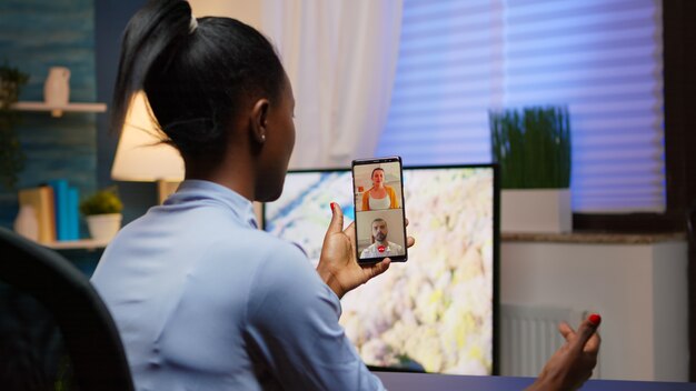 Black young lady discussing on webcam with family using smartphone taking a break after working overtime at home. Freelancer working in personal workplace having video meeting.