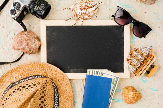 Blackboard near hat with toy boat and money among seashells and camera