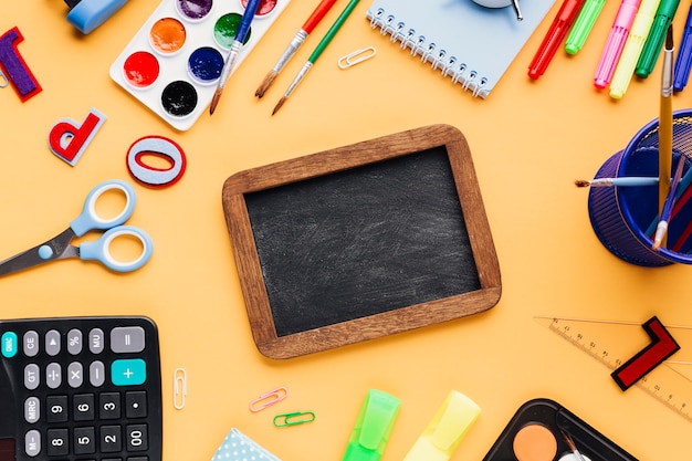 Blank blackboard surrounded with school supplies scattered on yellow desk