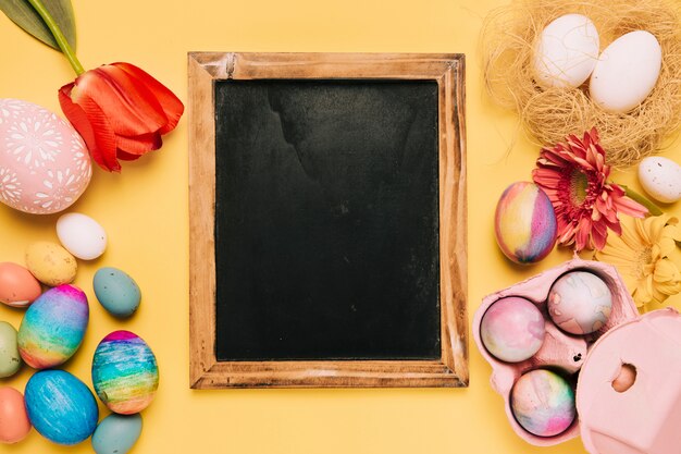 Blank blackboard with fresh flowers and decorated easter eggs on yellow backdrop