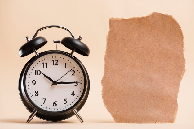 Blank brown torn paper near the black alarm clock against colored backdrop