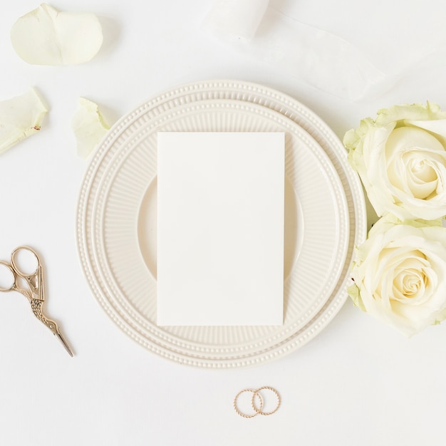 Blank card on ceramic plate with roses; scissor and wedding rings on white background