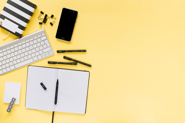 Blank diary with pens; bulldog paper clips; keyboard and cellphone on yellow background