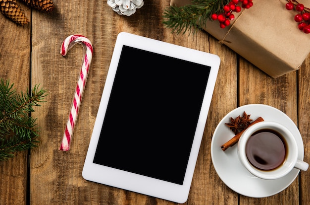 Blank empty screen of tablet on the wooden wall with colorful holiday's decoration, tea and gifts.