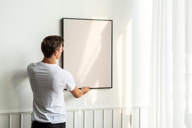 Free photo blank frame being hung by a young man on a white minimal wall