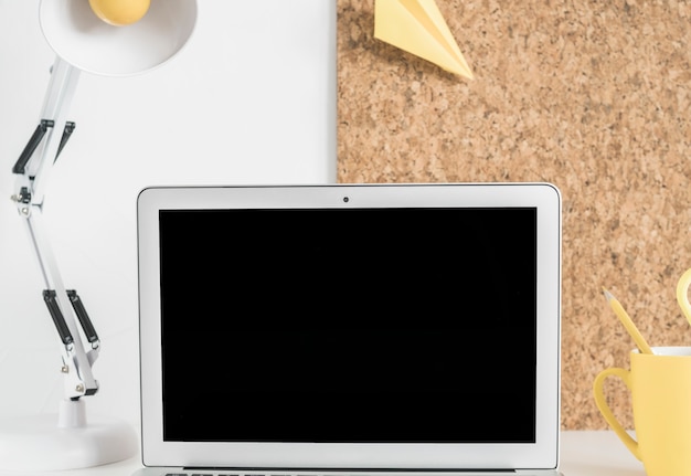 Blank laptop screen on desk with lamp and cork board