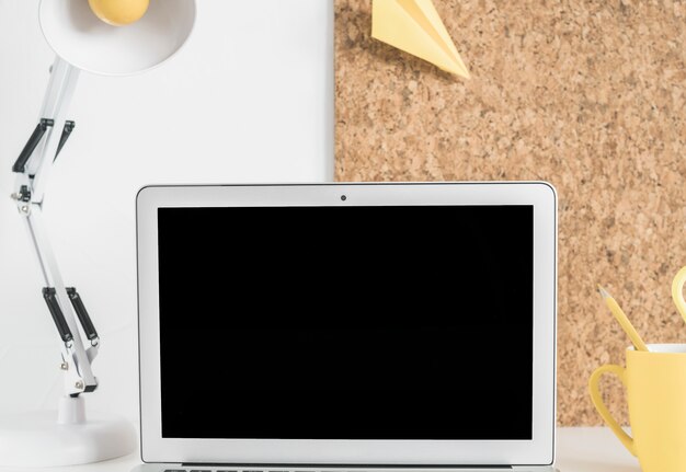 Blank laptop screen on desk with lamp and cork board