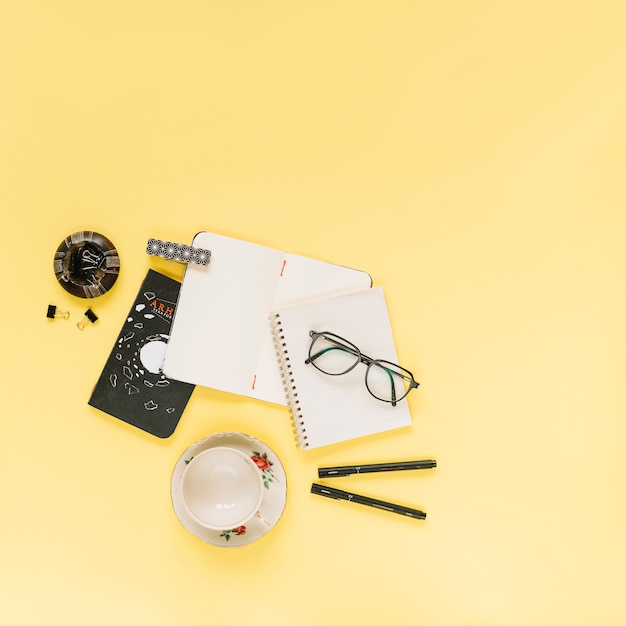 Blank notebooks; eyeglasses and pen with an empty cup on yellow background