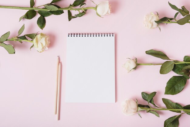 Blank notepad with two colored pencils surrounded with roses on pink backdrop