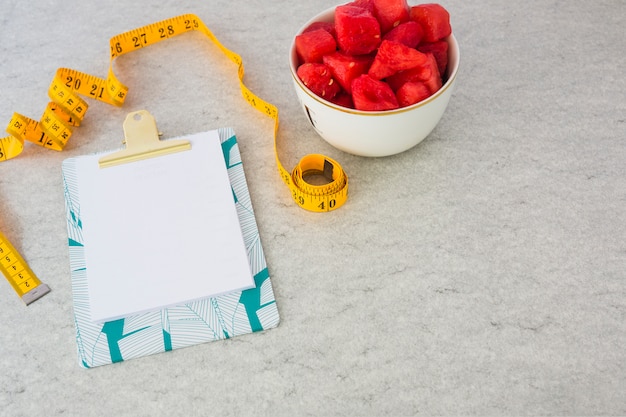 Blank paper attached on clipboard with measuring tape and watermelon cubes in the bowl