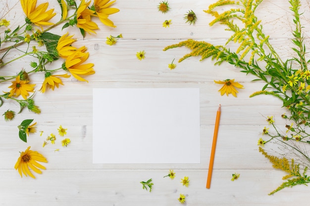 A blank paper beside a yellow pencil on a wooden surface with yellow-petaled flowers