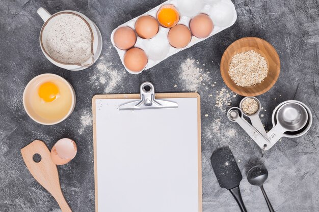 Blank paper on clipboard with baking ingredients over concrete backdrop