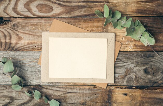 Blank paper and envelope on wooden background with eucalyptus twigs flat lay