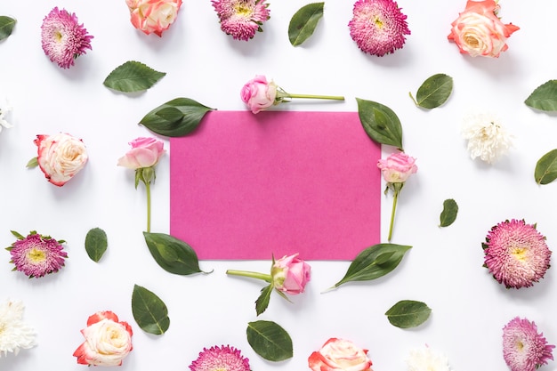 Blank pink paper surrounded by green leaves and flowers on white surface