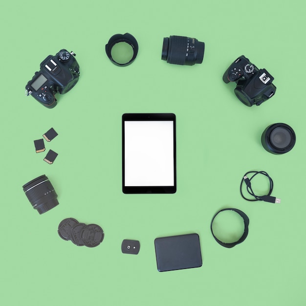 Blank screen digital tablet surrounded by professional digital camera and accessories over green background