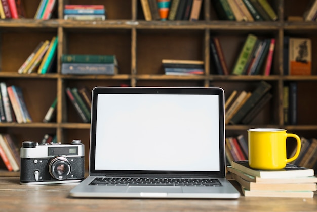 Blank screen laptop with retro camera; yellow coffee mug; stacked book on table in library room