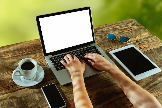 Blank screens of laptop and smartphone on a wooden table outdoors with nature on wall Coffee nearby. Concept of creative workplace, business, freelance. Copyspace. Hands typing text.