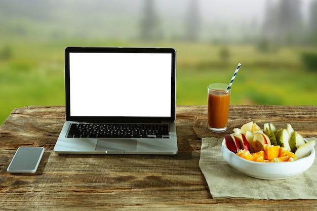 Blank screens of laptop and smartphone on a wooden table outdoors with nature on wall fruits and fresh juice nearby. concept of creative workplace, business, freelance. copyspace.