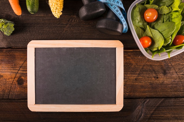 Blank slate with vegetables; dumbbell and measuring tape on wooden surface