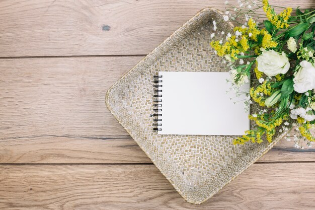 Blank spiral notepad and fresh flower bouquet in the basket on wooden desk