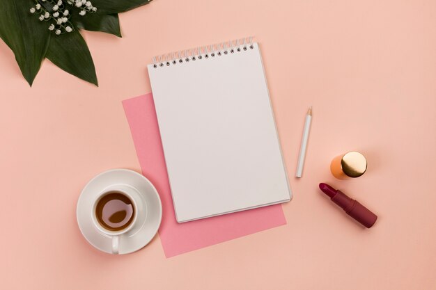 Blank spiral notepad,pencil,lipstick,coffee cup and leaves on peach background