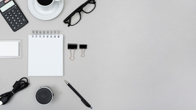 Blank spiral notepad surrounded with calculator; tea cup; paper clip; speaker; pen; cable and eye glasses over grey background