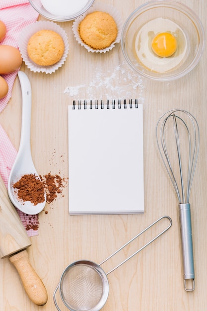 卵黄と空白のスパイラルメモ帳。カップケーキチョコレートパウダーと木製の机の上のツール