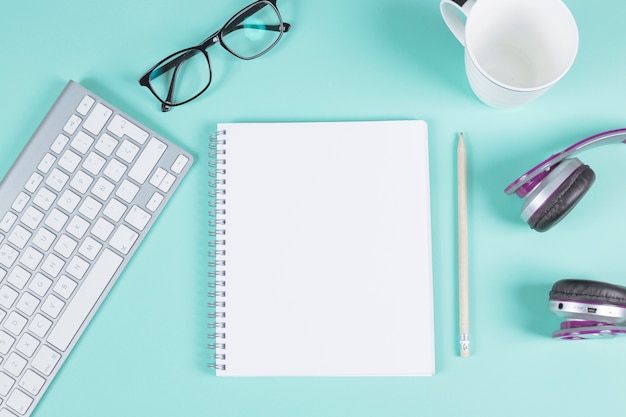 Blank spiral notepad with keyboard; eyeglasses; cup; headphone and pencil on turquoise background