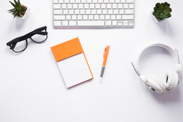 Blank sticky notepad; eyeglasses; pen; cactus plant; headphone and keyboard on white desk