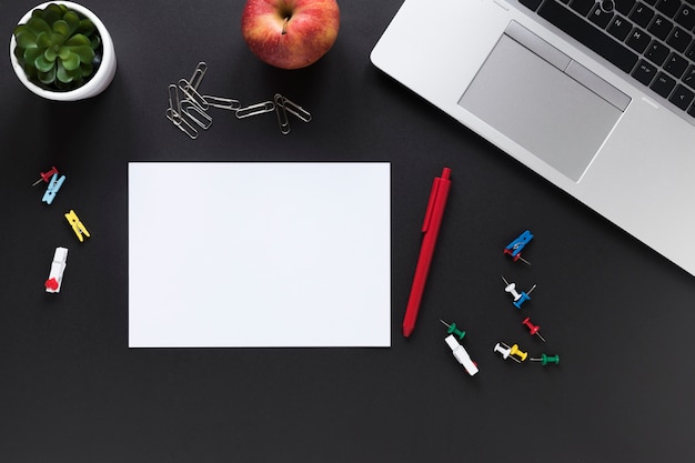 Blank white card paper with pen; apple; colorful office stationeries and laptop on black background