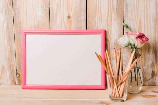 Blank white frame with pink border and colored pencils in glass on wooden desk