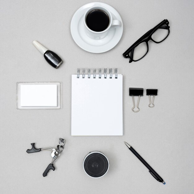 Blank white notepad surrounded by coffee cup; nail varnish; eyelash curler; speaker; pen and paper clips above gray background