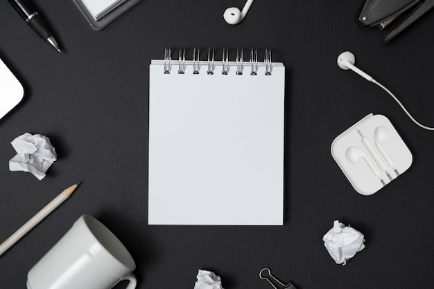 Blank white notepad surrounded by empty cup; crumpled paper; pen; earphone over black backdrop