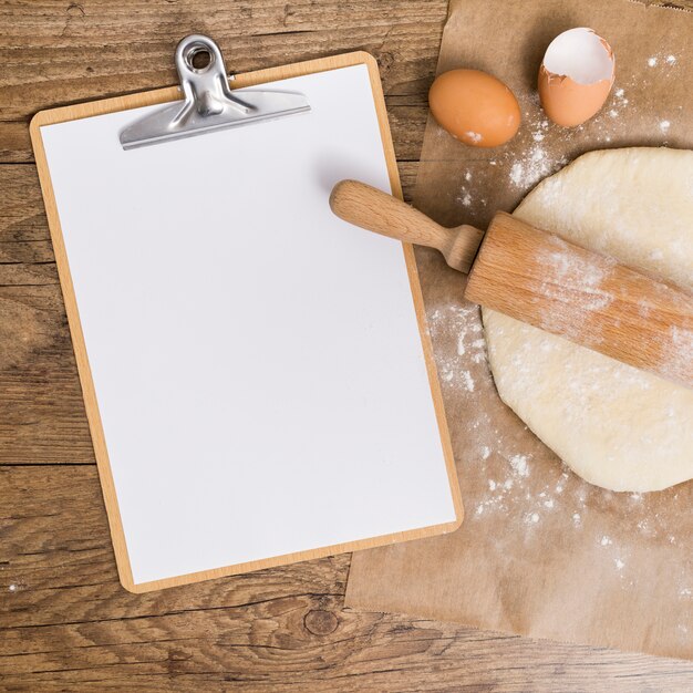 Blank white paper on clipboard; flat dough and egg shells on parchment paper over the wooden table