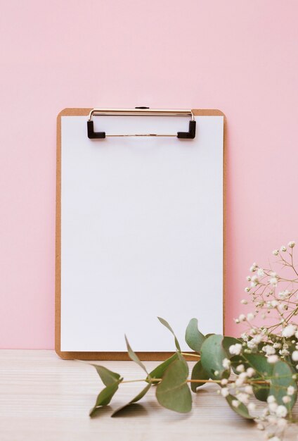 Blank white paper on clipboard with leaves and baby's-breath flowers on wooden desk against pink background