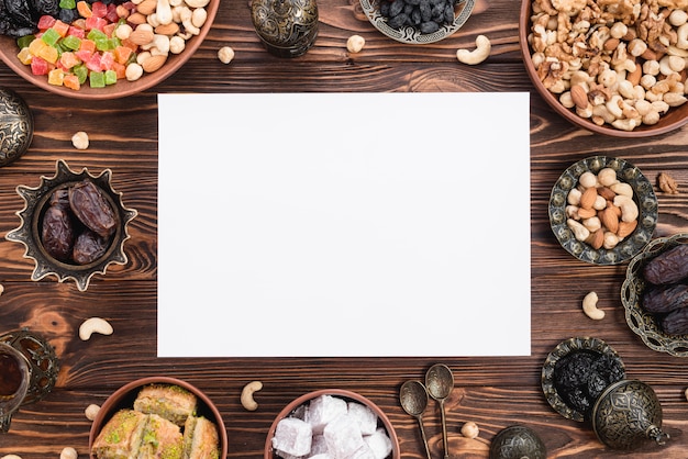 Blank white paper surrounded with mix dried fruits; dates; lukum; baklava and nuts on wooden desk for ramadan