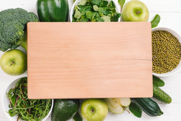 Blank wooden chopping board over the green vegetables and fruits