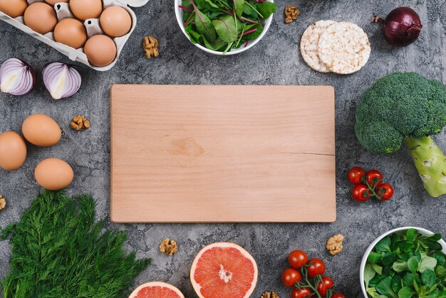 Пустая деревянная разделочная доска с сырыми овощами и воздушный рисовый пирог на бетонном фоне