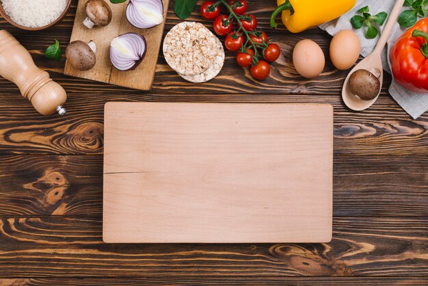 Blank wooden chopping board with raw vegetables and puffed rice cake over wooden desk