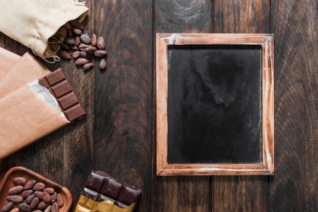 Blank wooden slate with cocoa beans and chocolate bars on wooden table