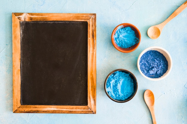 Blank wooden slate with holi colored bowls and wooden spoon on colored background