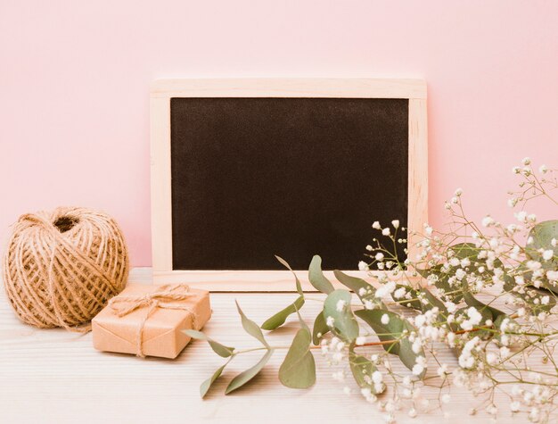 Blank wooden slate with spool; gift box and baby's-breath flowers on wooden desk against pink background