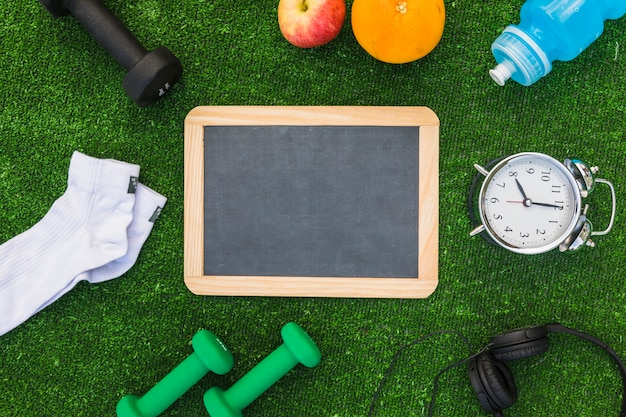 Blank wooden slate with sport equipments and healthy fruits on green turf